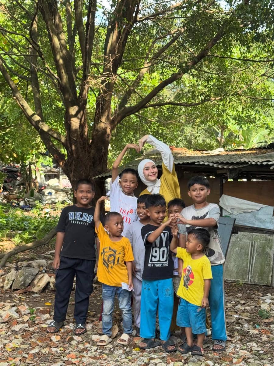 Seorang wanita berhijab berpose bersama sekelompok anak-anak di area terbuka dengan latar pepohonan dan bangunan sederhana.
