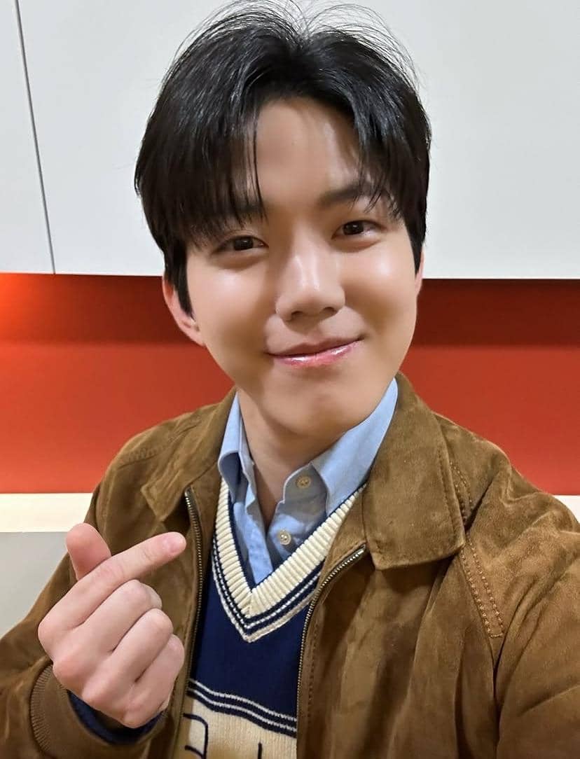 Seorang pria berambut hitam mengenakan jaket cokelat dan kemeja biru muda tersenyum sambil membuat gestur hati dengan jarinya.