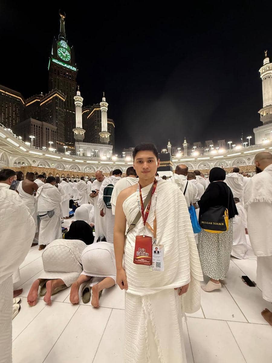 Seorang jamaah mengenakan pakaian ihram berdiri di area Masjidil Haram pada malam hari dengan latar belakang Menara Abraj Al Bait.