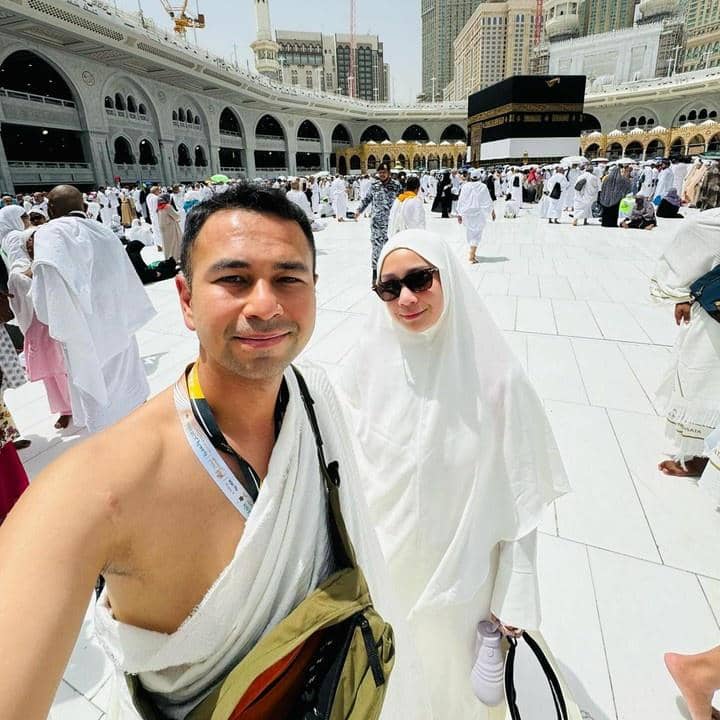Sepasang jamaah mengenakan pakaian ihram dan mukena putih berpose di area Masjidil Haram dengan latar belakang Ka'bah di Makkah.