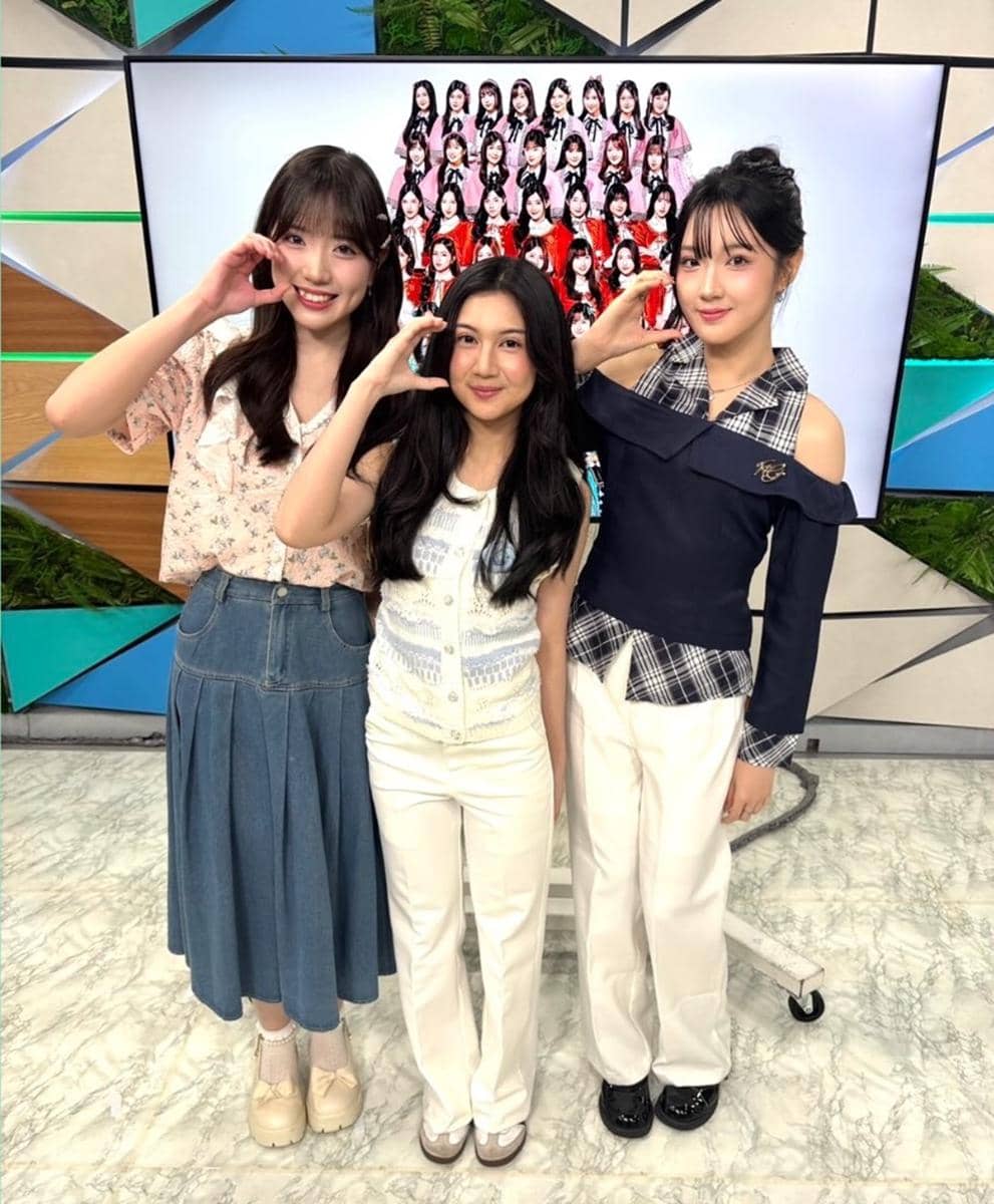 Tiga perempuan berdiri berdampingan di studio TV Jepang dengan latar layar menampilkan grup besar perempuan berpakaian merah.