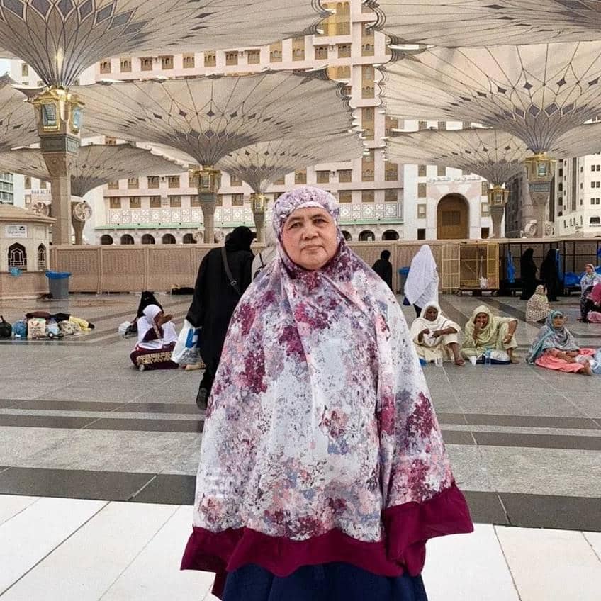 Seorang perempuan mengenakan mukena bermotif bunga berwarna ungu dan putih berdiri di area masjid dengan payung besar terbuka di latar belakang.