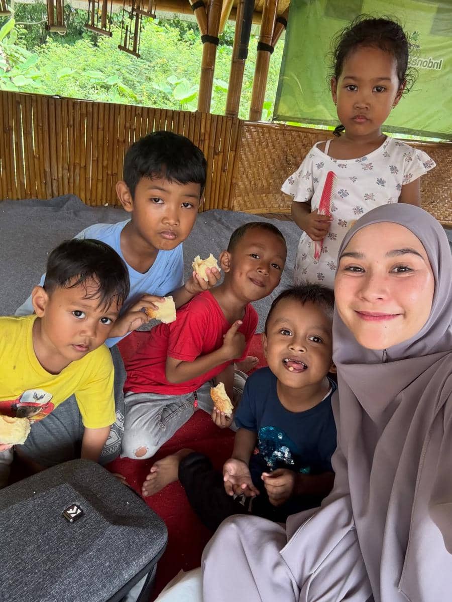 Seorang perempuan berhijab tersenyum bersama lima anak kecil yang sedang duduk dan menikmati roti di ruangan bambu terbuka.