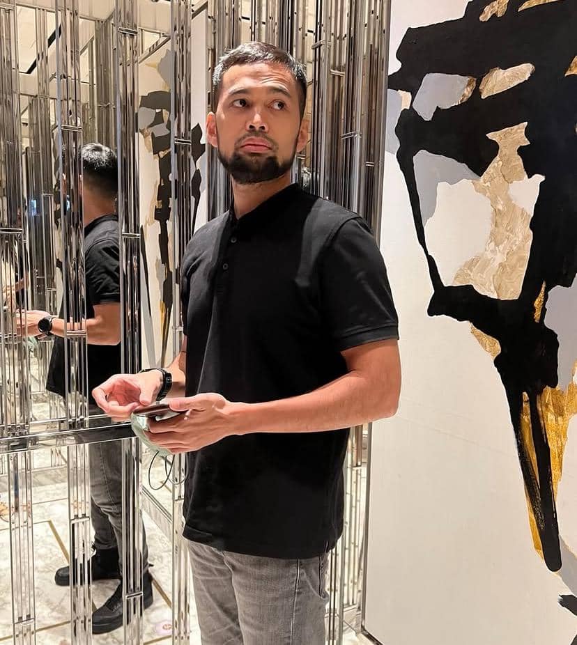 Seorang pria berpenampilan kasual mengenakan kaus hitam dan celana abu-abu berdiri di ruangan dengan dinding cermin dan dekorasi abstrak.