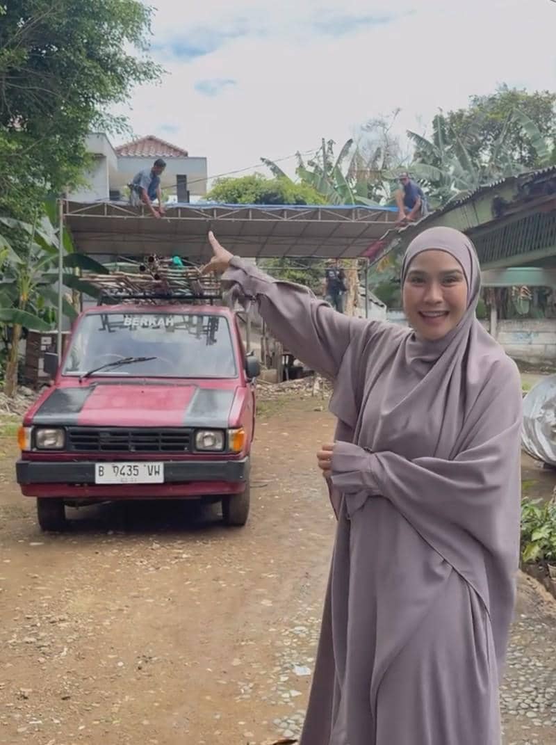 Seorang perempuan berhijab abu-abu tersenyum sambil menunjuk ke arah pekerja yang sedang memasang tenda di atas mobil pickup merah.