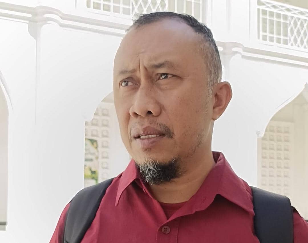 Seorang pria mengenakan kemeja merah dan tas punggung hitam berdiri di area dengan latar belakang arsitektur putih bercorak lengkung.