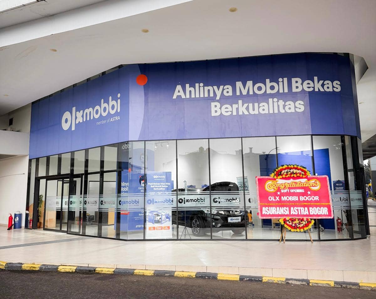 Tampak depan outlet OLXMobbi Bogor dengan kaca besar, papan nama biru, dan karangan bunga ucapan pembukaan di sisi kanan.