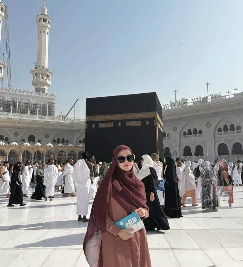 Seorang perempuan berhijab berwarna marun memegang buku di area Masjidil Haram dengan latar belakang Ka'bah dan jamaah yang beribadah.