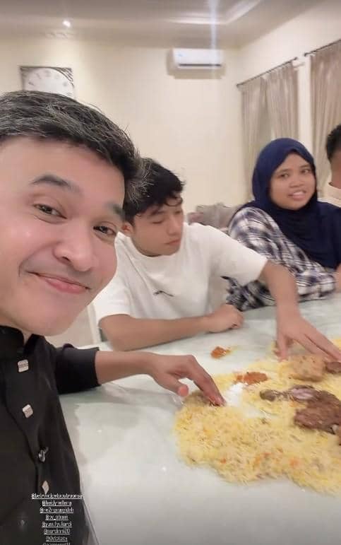 Seorang pria tersenyum bersama dua orang lainnya sedang menikmati hidangan nasi di meja saat berbuka puasa di dalam ruangan.