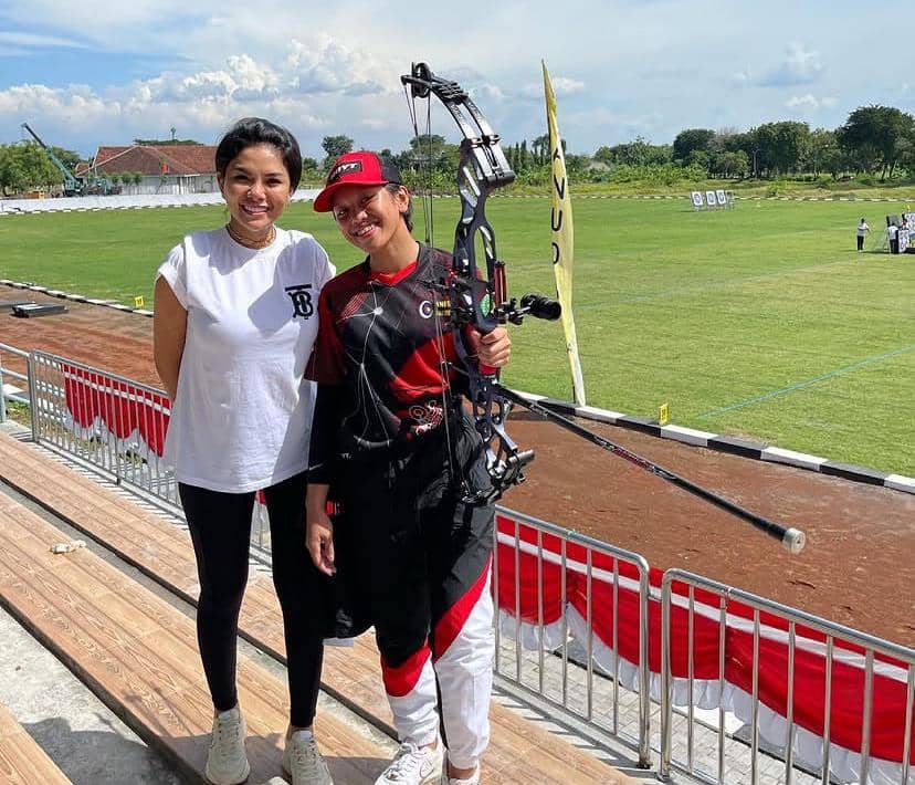 Dua perempuan berpose di tribun lapangan panahan, satu mengenakan seragam olahraga dan memegang busur panah, satunya mengenakan kaus putih.