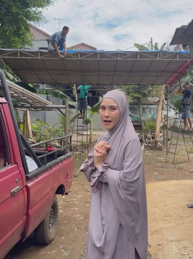 Seorang perempuan berhijab abu-abu berdiri di dekat mobil pikap merah, sementara beberapa pria memasang tenda di latar belakang.