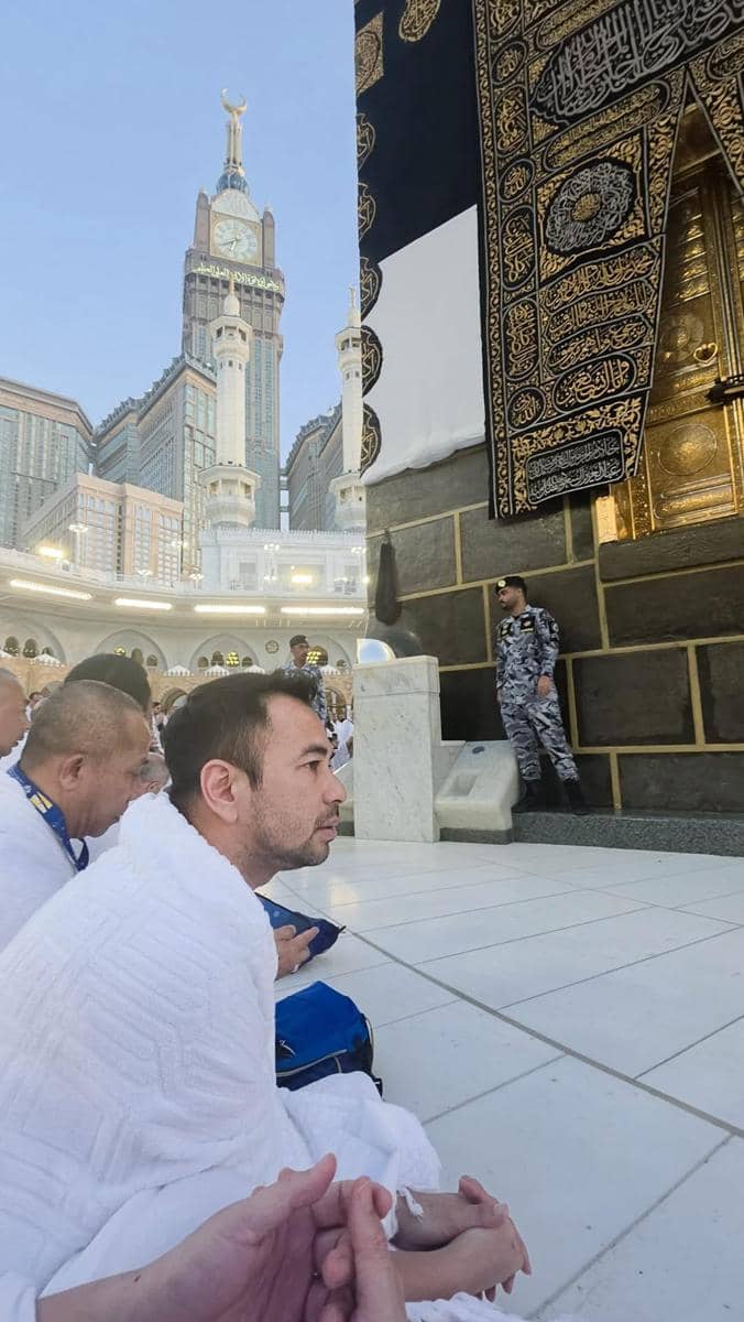 Seorang jamaah pria mengenakan kain ihram duduk berdoa di dekat Ka'bah di Masjidil Haram, dengan latar menara jam Makkah.