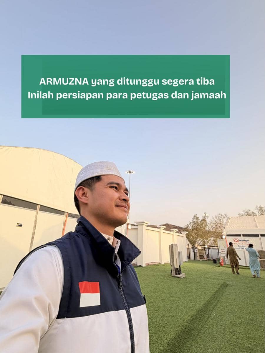 Seorang pria mengenakan rompi dengan bendera Indonesia berdiri di area tenda haji, tampak langit cerah dan beberapa orang berjalan di kejauhan.