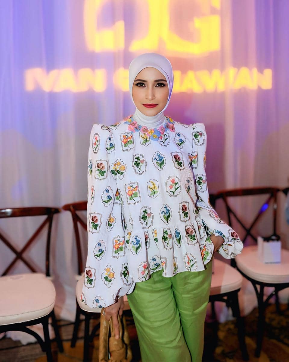 Seorang wanita berhijab mengenakan blus putih bermotif bunga warna-warni dan celana hijau muda, berpose di acara dengan latar bertulisan Ivan Gunawan.