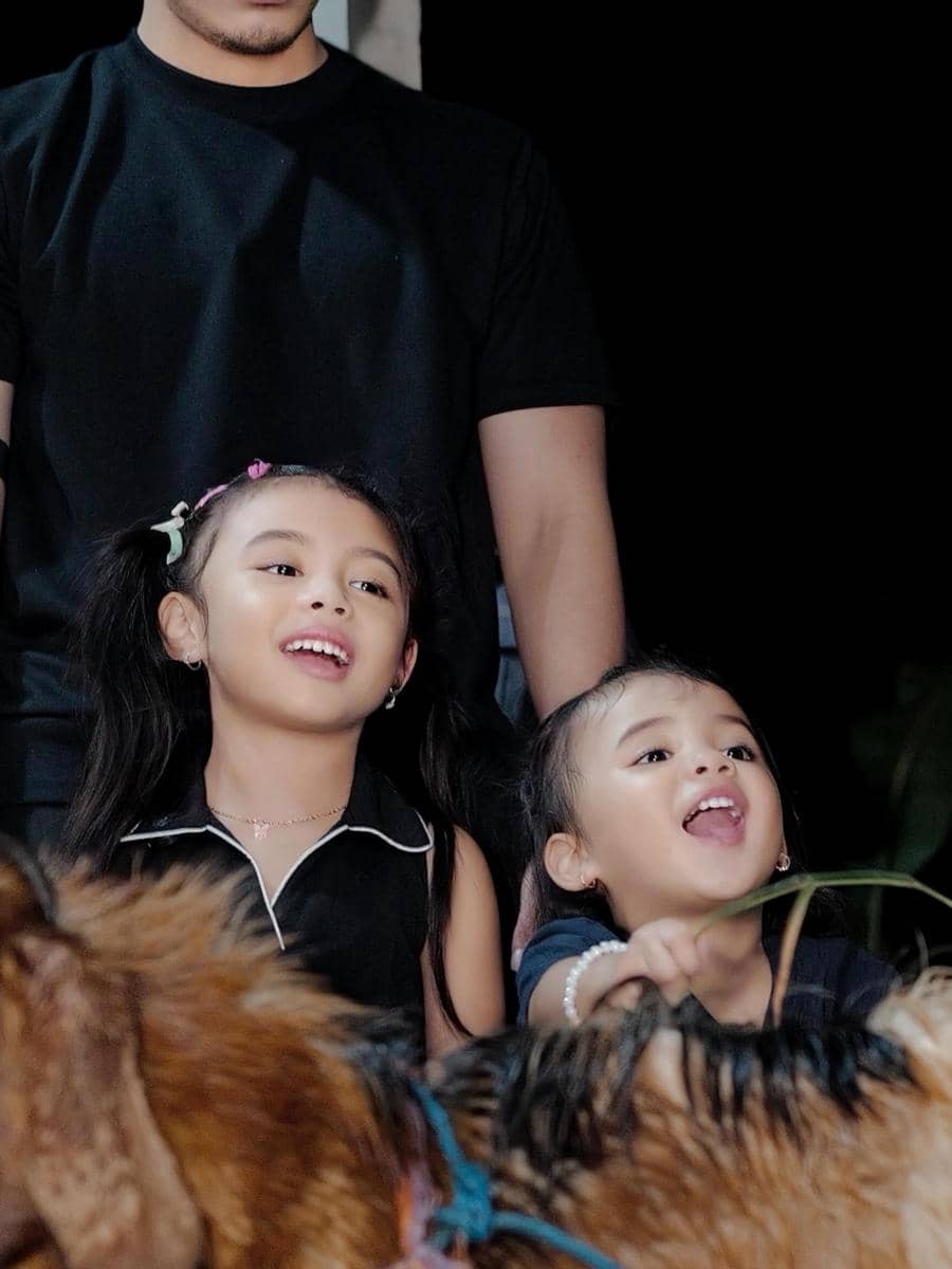 Dua anak kecil tersenyum gembira sambil melihat hewan kurban di depan mereka, ditemani seorang pria berpakaian hitam.