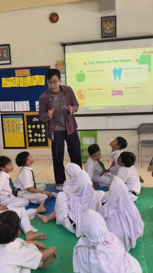 Seorang pria berdiri di depan murid-murid SD yang duduk di lantai kelas sambil menjelaskan materi kesehatan dengan presentasi di layar proyektor.