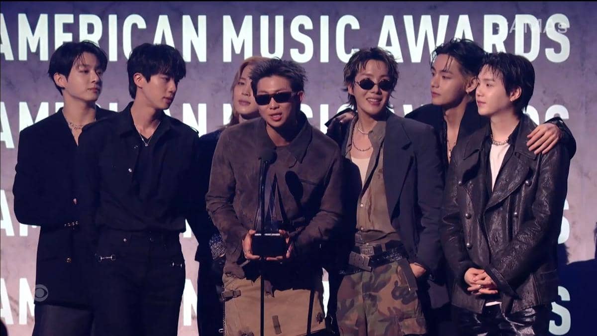 Grup musik BTS berdiri di panggung American Music Awards 2026 sambil memegang trofi penghargaan dan berbicara di depan mikrofon.