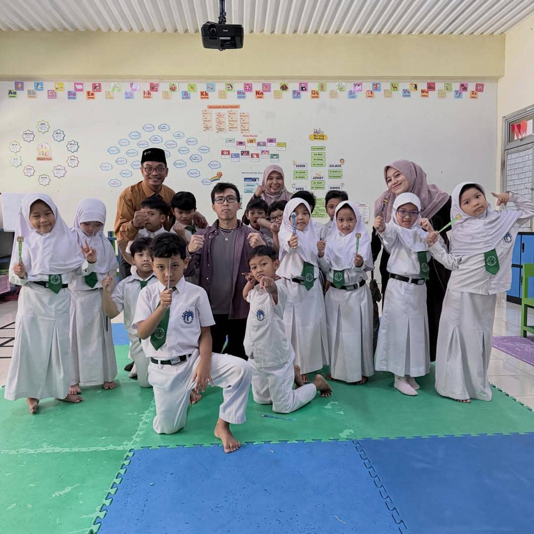 Seorang pria berdiri bersama murid-murid kelas 1 SD dan beberapa guru di ruang kelas yang penuh hiasan edukatif di dinding.