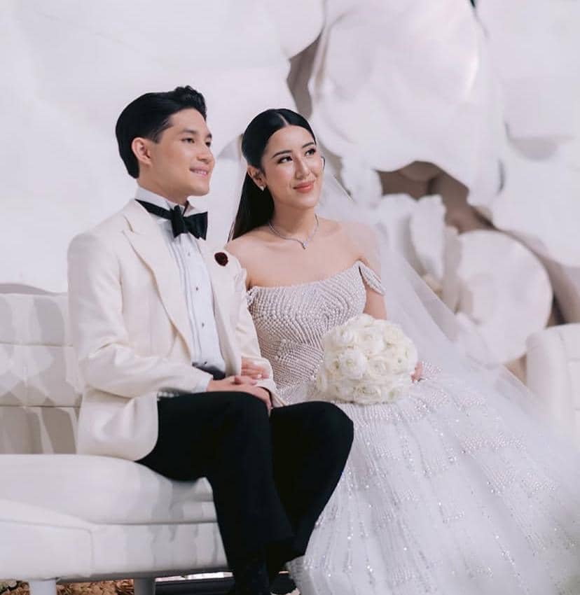 Sepasang pengantin duduk berdampingan mengenakan busana pernikahan elegan, sang pria memakai jas putih dan wanita mengenakan gaun putih berpayet.
