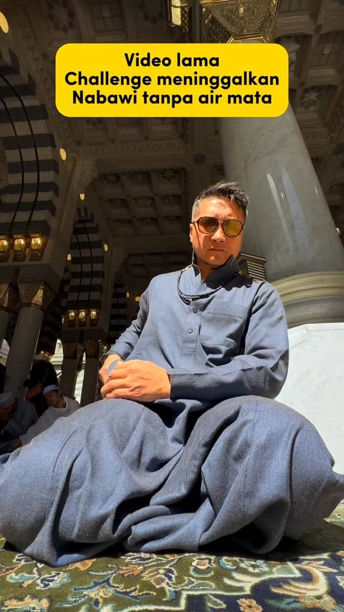 Seorang pria mengenakan gamis biru dan kacamata hitam duduk bersila di area Masjid Nabawi dengan cahaya matahari menyinari wajahnya.