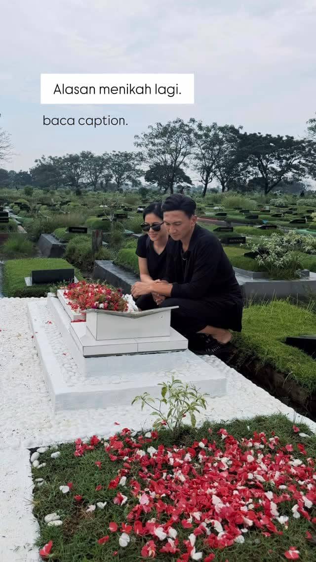 Seorang pria dan wanita berpakaian hitam duduk di samping makam yang dihiasi bunga tabur merah putih di area pemakaman hijau.