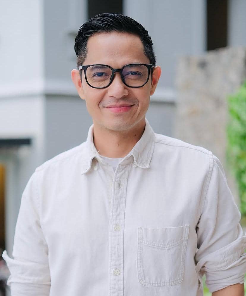 Seorang pria berkacamata mengenakan kemeja putih tersenyum di luar ruangan dengan latar bangunan dan tanaman hijau.