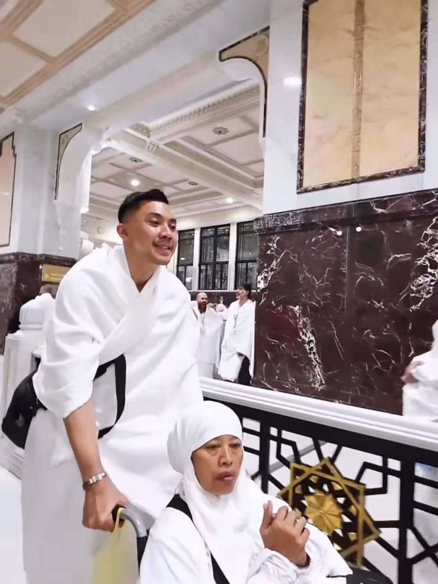 Seorang pria berpakaian ihram mendorong kursi roda yang diduduki seorang wanita berhijab putih di area dalam masjid dengan dinding marmer.