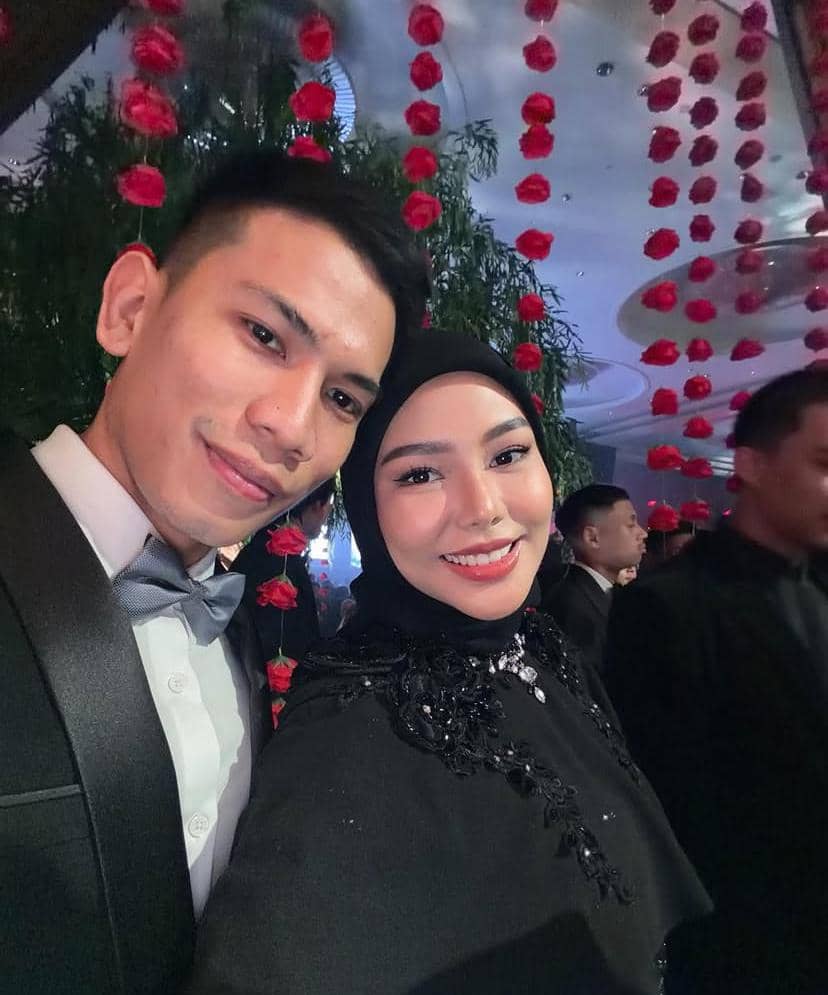 Sepasang pria dan wanita berpakaian formal berpose bersama di acara malam dengan dekorasi bunga mawar merah menggantung di latar belakang.