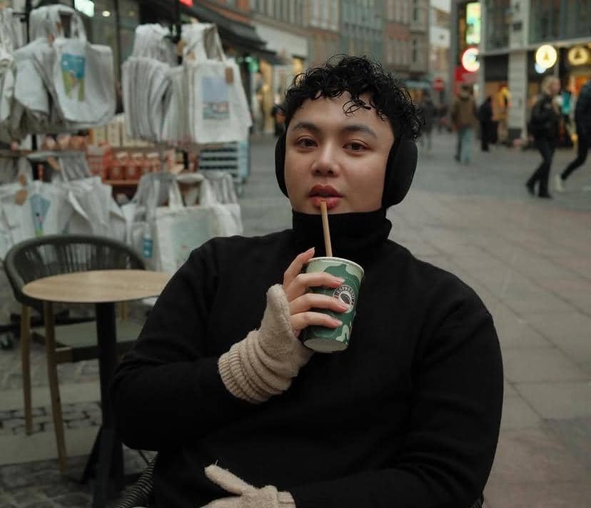 Seseorang mengenakan sweater hitam dan penutup telinga sambil memegang minuman di area luar ruangan dengan latar toko dan tas kain.