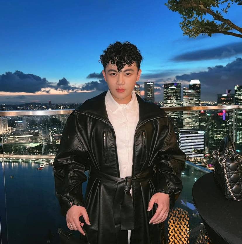 Seorang pria berjaket kulit hitam dan kemeja putih berpose di balkon gedung tinggi dengan latar pemandangan kota saat senja.