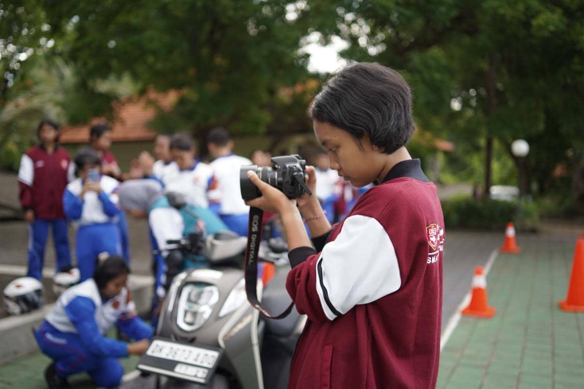 Seorang pelajar mengenakan jaket marun sedang memegang kamera dan memotret di area luar bersama teman-temannya.