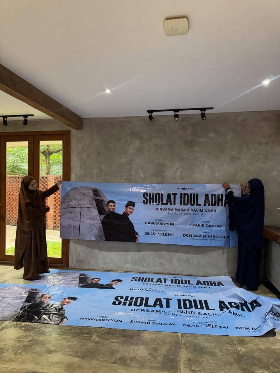 Dua perempuan berhijab sedang memasang spanduk acara salat Iduladha 2026 di dalam ruangan dengan dinding abu-abu dan jendela kayu besar.