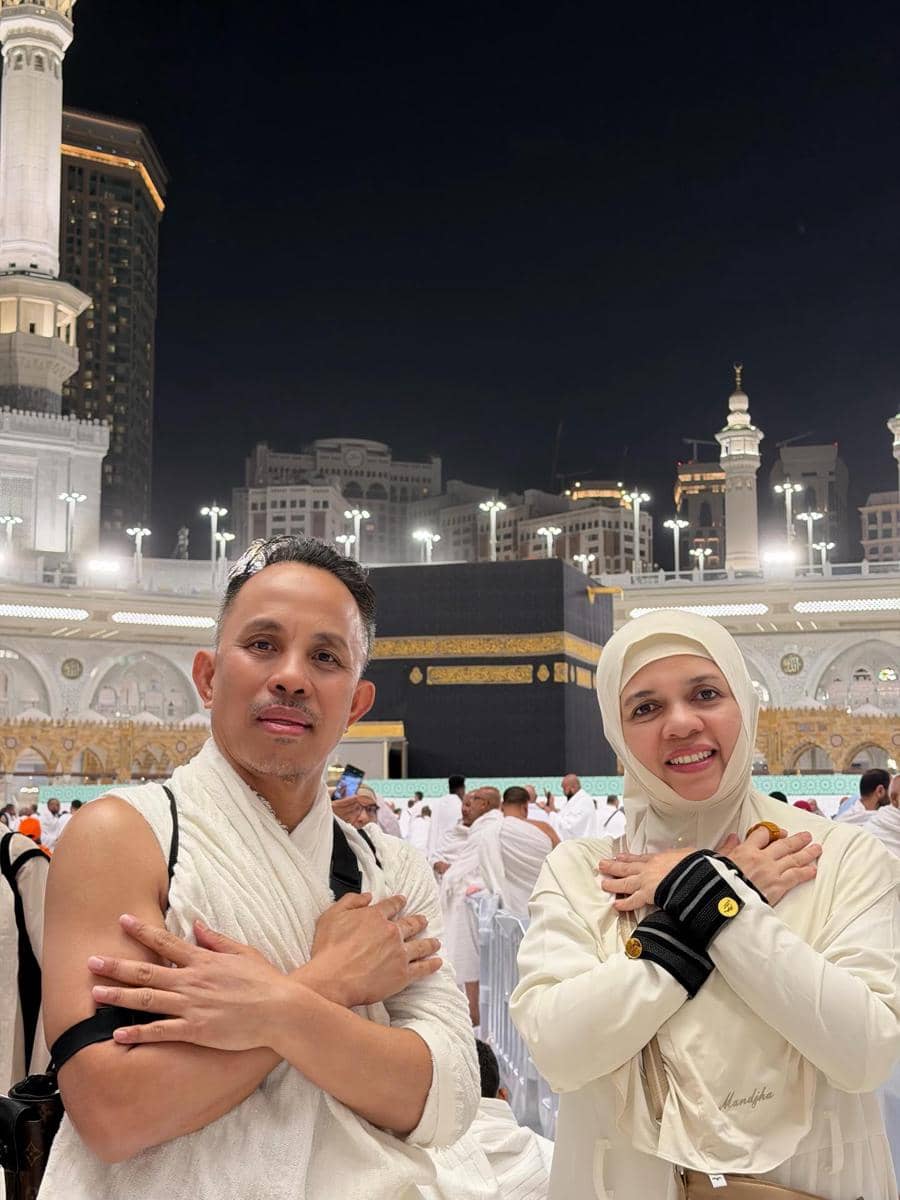 Sepasang jamaah mengenakan pakaian ihram berpose di depan Ka'bah pada malam hari dengan latar Masjidil Haram yang terang.