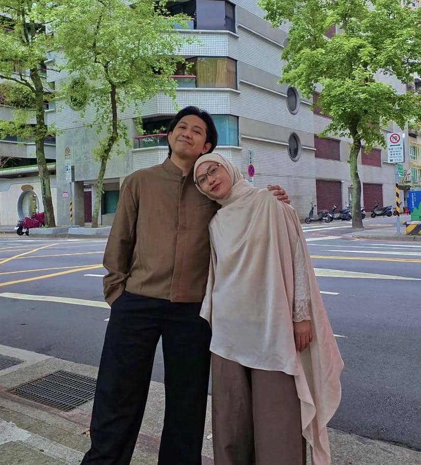 Seorang pria dan wanita berpose bersama di tepi jalan kota dengan latar gedung bertingkat dan pepohonan rindang di sekitarnya.