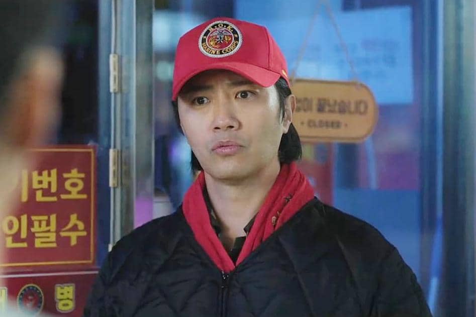 Seorang pria mengenakan topi merah dan jaket hitam berdiri di depan pintu kaca dalam adegan drama Korea The Legend of Kitchen Soldier.