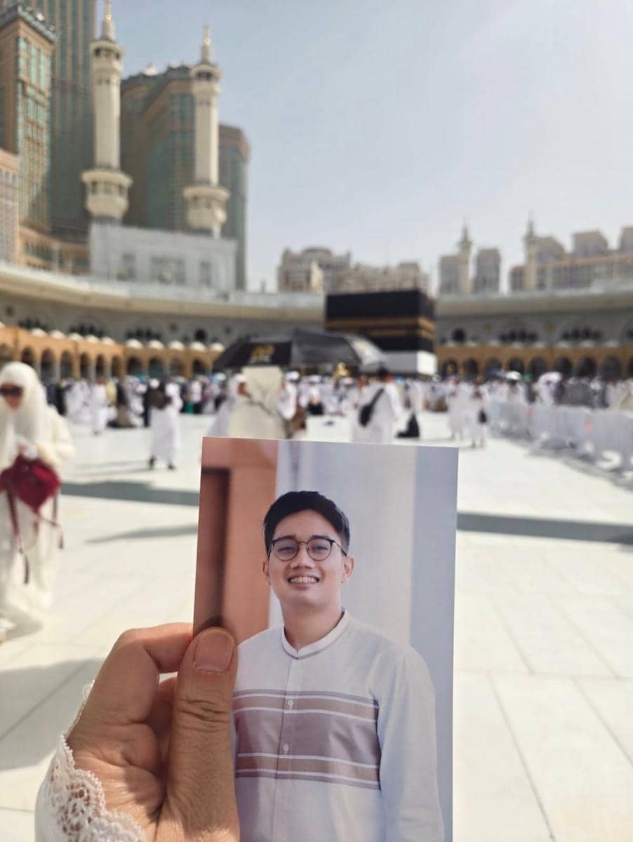 Seseorang memegang foto pria muda tersenyum di depan Ka'bah di Masjidil Haram, dengan latar jamaah dan bangunan tinggi di Mekkah.