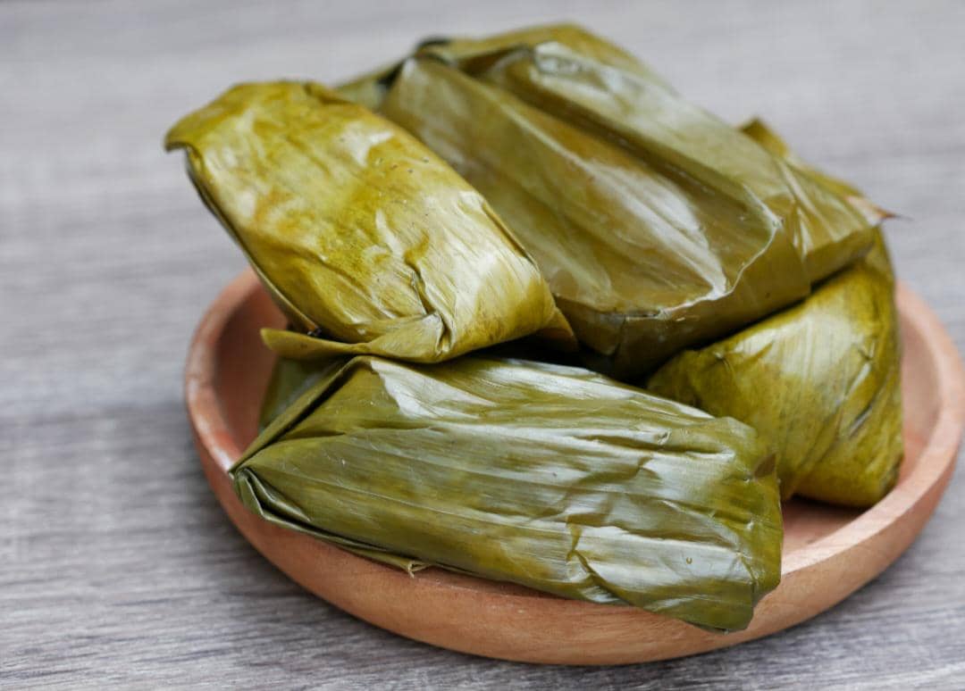 Beberapa potong lontong dibungkus daun pisang disajikan di atas piring kayu di atas meja berwarna abu-abu.