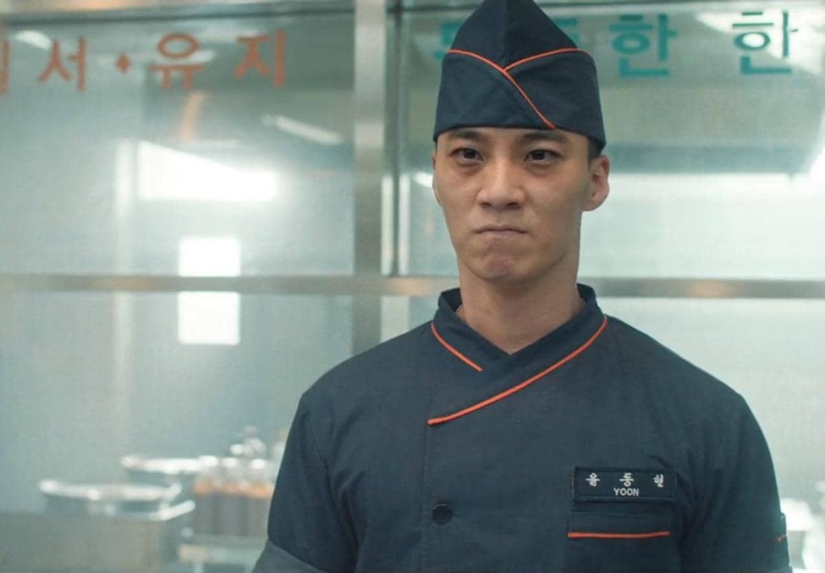 Seorang pria mengenakan seragam dapur berwarna gelap dengan garis oranye berdiri di dapur dalam adegan drama The Legend of Kitchen Soldier.