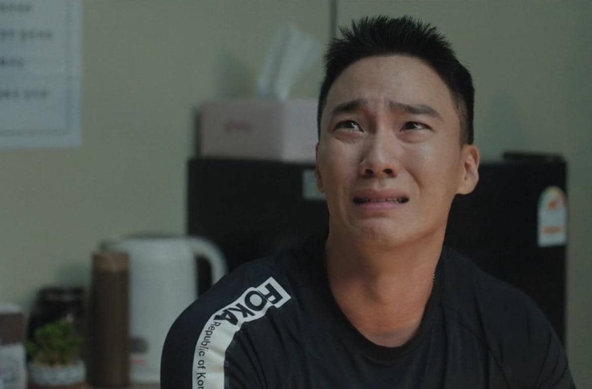 Seorang pria berseragam latihan militer dengan ekspresi sedih di dalam ruangan, adegan dari drama The Legend of Kitchen Soldier.