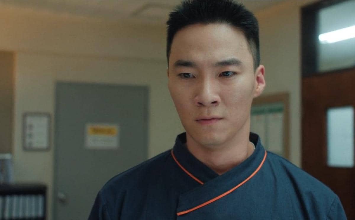 Seorang pria berambut pendek mengenakan seragam dapur berwarna gelap dengan garis oranye dalam adegan drama The Legend of Kitchen Soldier.