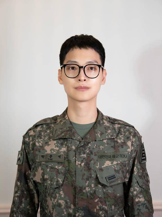 Seorang pria mengenakan seragam militer Korea Selatan lengkap dengan kacamata hitam berbingkai tebal, berdiri tegak di latar polos.