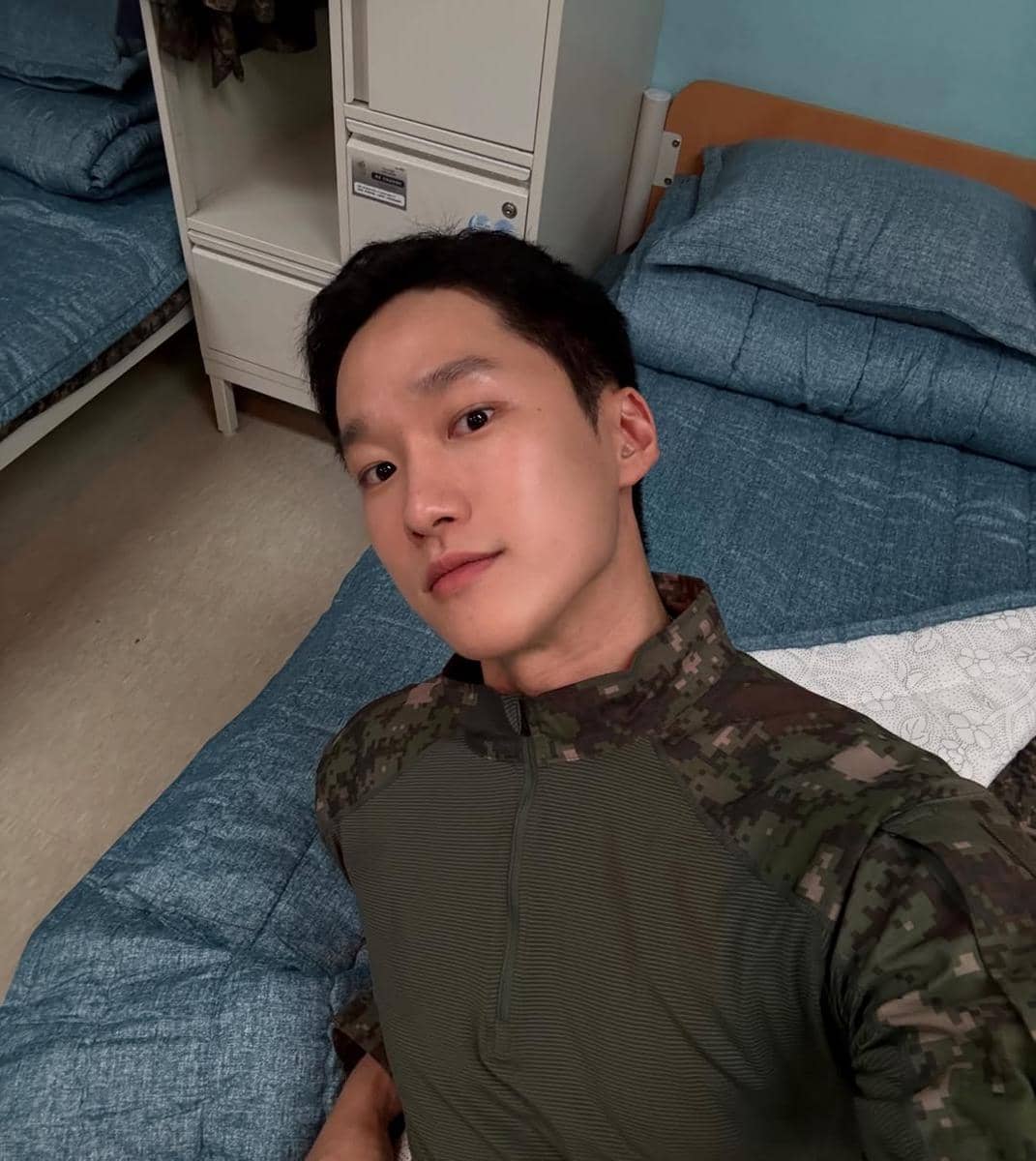 Seorang pria mengenakan seragam militer berpose selfie di kamar dengan tempat tidur berseprai biru dan lemari kecil di belakangnya.