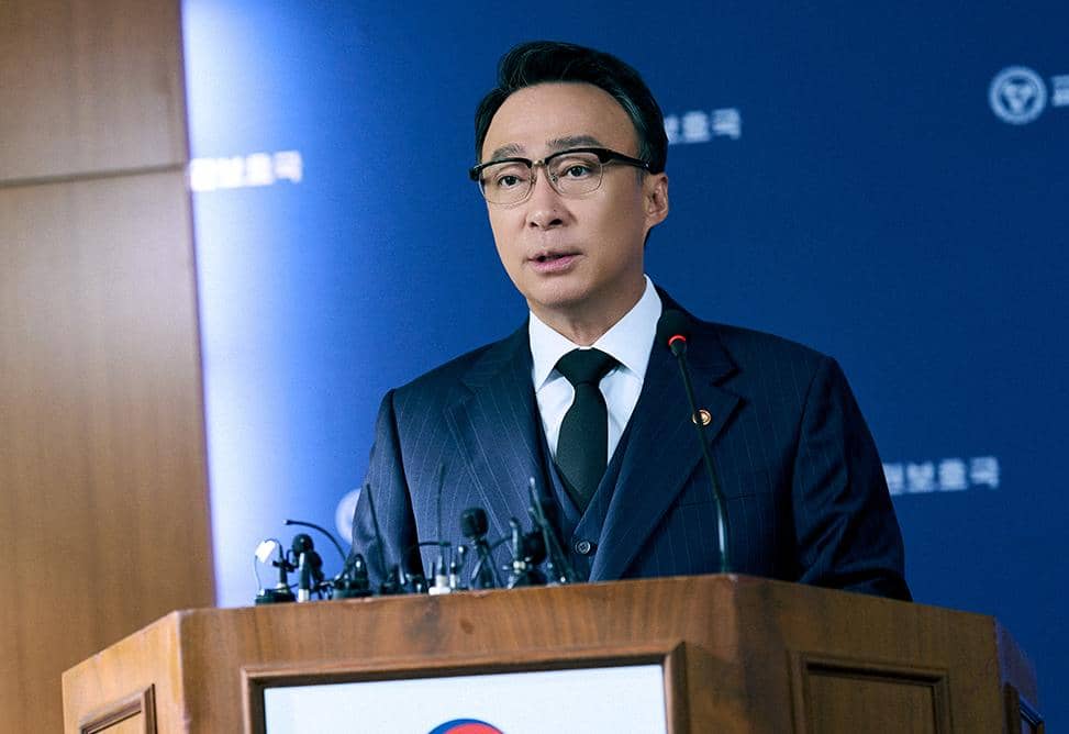 Seorang pria mengenakan setelan jas gelap dan kacamata sedang berbicara di podium dengan mikrofon di depannya dalam adegan drama Korea Teach You a Lesson.