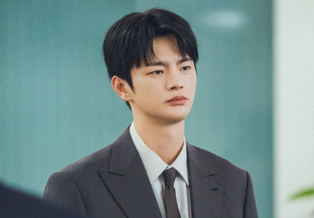 Seorang pria berjas abu-abu dan dasi cokelat berdiri di dalam ruangan dengan ekspresi serius, diambil dari drama Korea See You at Work Tomorrow.