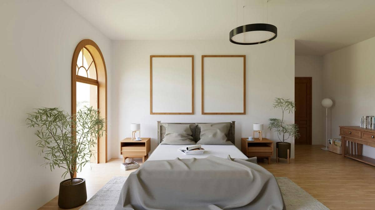 Kamar tidur luas dengan jendela lengkung bergaya Mediterania, tempat tidur queen berselimut abu-abu, dua nakas kayu, tanaman hias hijau, lampu gantung lingkaran hitam, dan dua bingkai foto kosong di dinding.