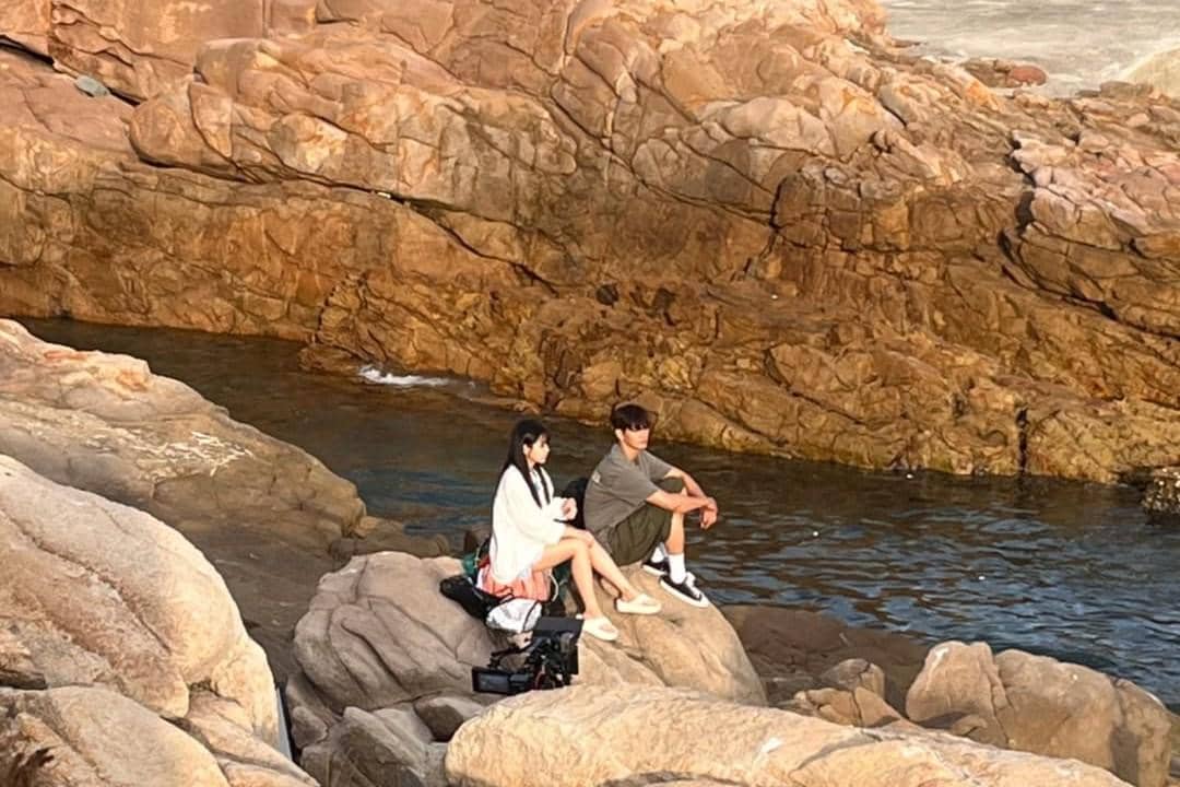 Dua orang duduk di atas batu besar di tepi sungai dangkal dengan latar tebing batu cokelat dalam adegan drama Azure Spring.