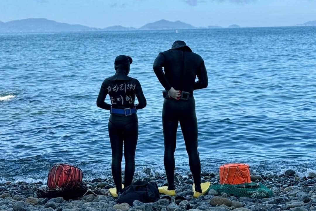 Dua penyelam mengenakan pakaian selam hitam berdiri di tepi pantai berbatu dengan lautan biru dan pulau di kejauhan.