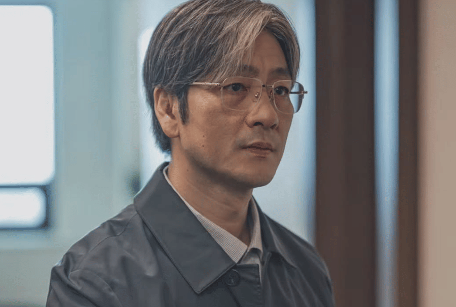 Seorang pria berambut abu-abu mengenakan kacamata dan jaket abu-abu tampak serius dalam cuplikan drama Korea The Scarecrow.