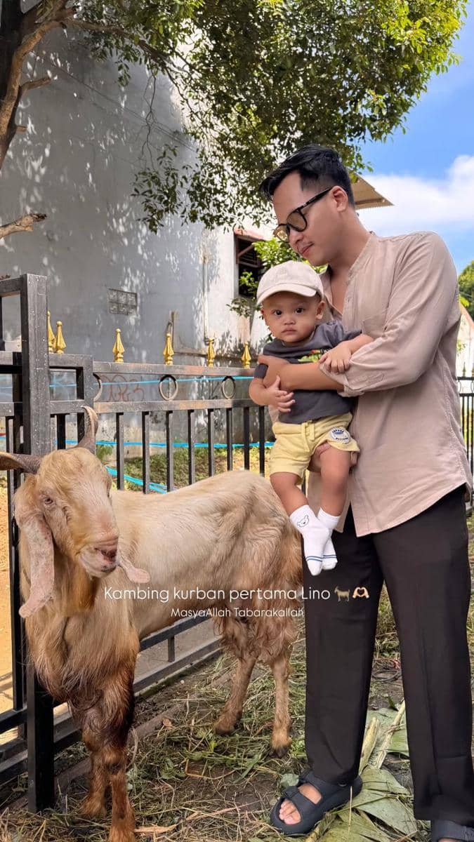 Seorang pria memegang bayi kecil di dekat kambing kurban berwarna cokelat muda di area berpagar dengan latar pepohonan dan bangunan.