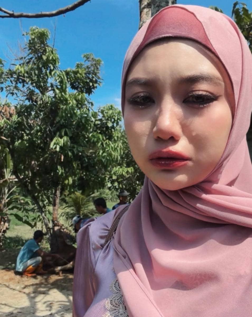 Perempuan berhijab warna merah muda tampak emosional di luar ruangan dengan latar beberapa orang sedang mempersiapkan hewan kurban.